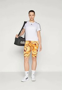 Sportos öltözék, amely fehér rövid ujjas pólóból, sárga mintás rövidnadrágból és fehér sportcipőből áll, egy fekete Adidas tornazsákkal kiegészítve.