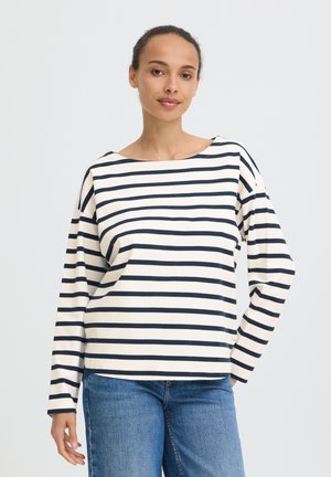 Femme portant une chemise blanche à manches longues avec des rayures horizontales noires et un jean bleu, debout avec une main sur la hanche.