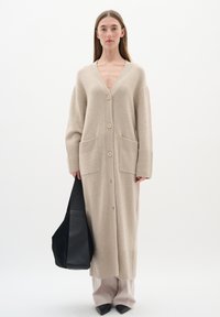 Beige lång cardigan i mjukt stickat material med V-ringning, fem knappar, ribbade ärmmuddar, två framfickor och sidslitsar.