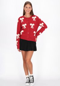 Fiatal nő mosolyog, piros pulóvert visel fehér masnikkal, fekete pliszírozott szoknyát, fehér zoknit és fekete magas szárú sportcipőt, kézzel a csípőjén áll.