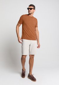 Bruine T-shirt met korte mouwen, lichtbeige korte broek en bruine schoenen met roze veters. Model staat en draagt een zwart zonnebril. Eenvoudig ontwerp, gladde textuur.