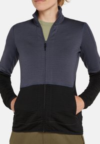 Chaqueta de forro polar con cierre frontal, con un diseño en bloques de color en gris oscuro y negro, mangas largas y bolsillos laterales. Textura elástica.