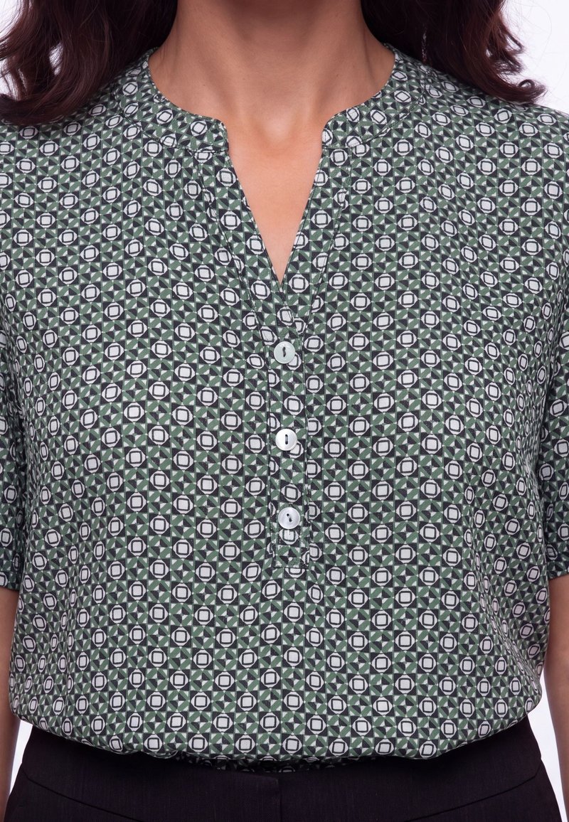 Femme portant une chemise à manches courtes boutonnée avec un motif géométrique vert, noir et blanc et un pantalon noir.