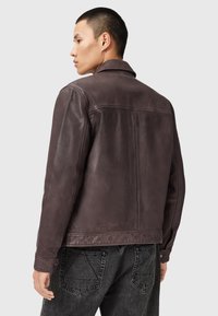 Jeune homme de profil portant une veste en cuir marron foncé et un jean noir sur un fond gris clair.