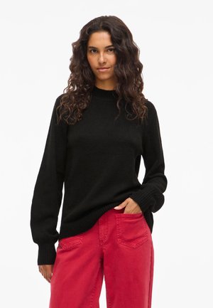 Woman with long curly hair wearing a black sweater and high-waisted red pants, standing with one hand in pocket against a white background.