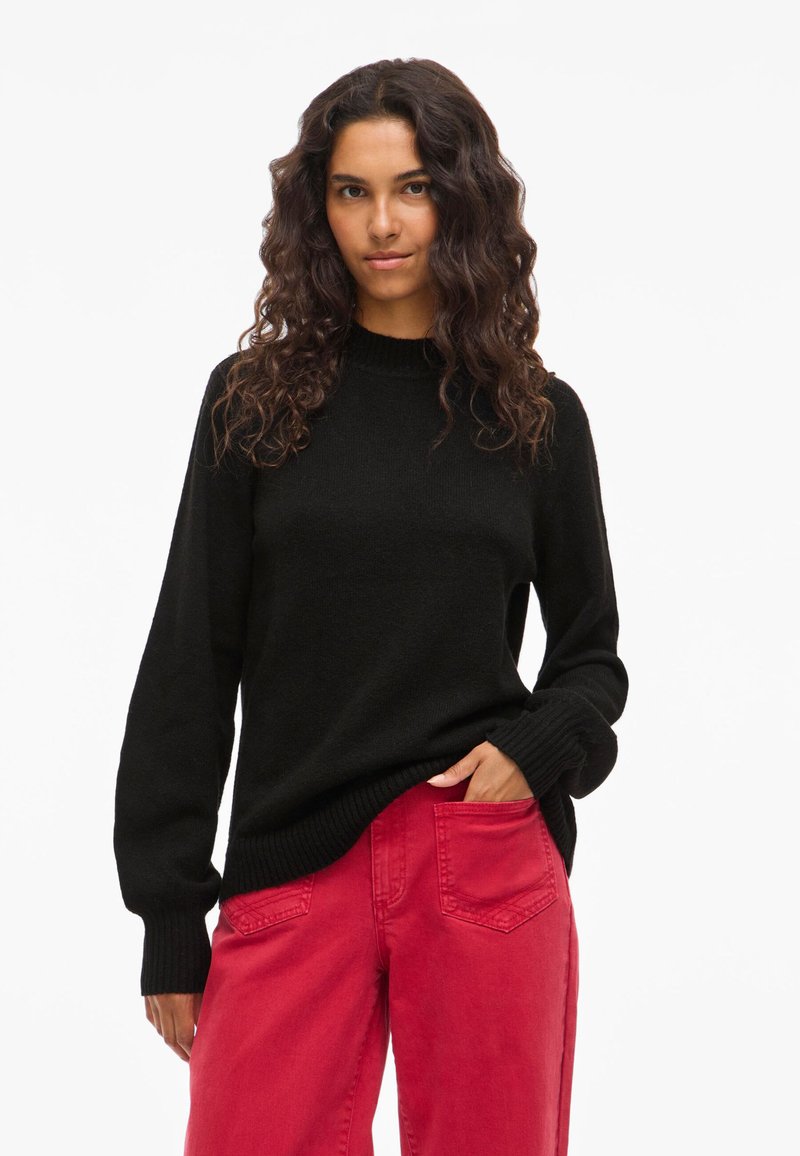 Γυναίκα με μακριά σγουρά μαλλιά, φορώντας ένα μαύρο πουλόβερ και κόκκινες παντελόνες ψηλής μέσης, στέκεται με το ένα χέρι στην τσέπη της, μπροστά σε λευκό φόντο.