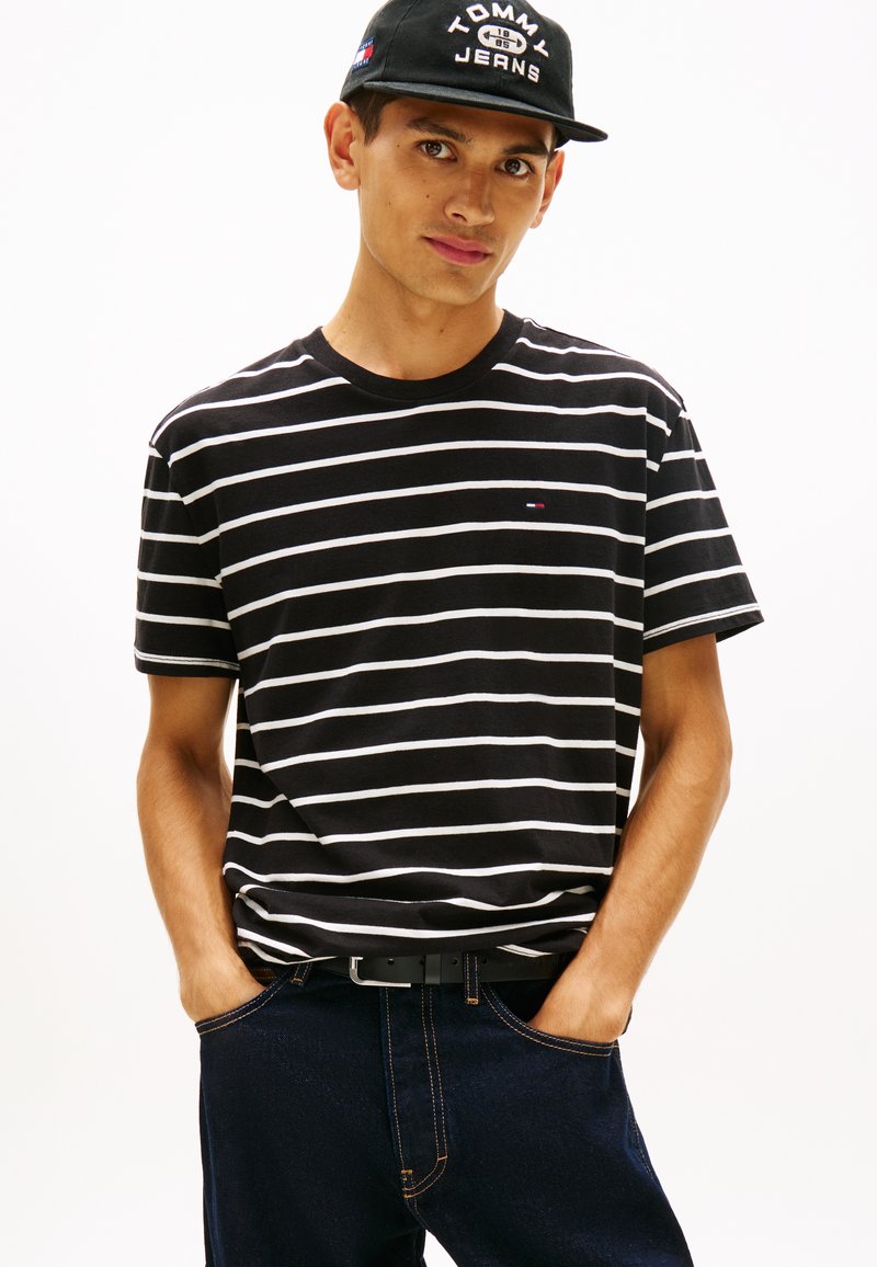 Νέος άντρας που φοράει μαύρο και άσπρο ριγέ μπλουζάκι, σκουρόχρωμο τζιν και μαύρο καπέλο με το λογότυπο "Tommy Jeans", με τα χέρια στις τσέπες, κοιτάζει μπροστά.