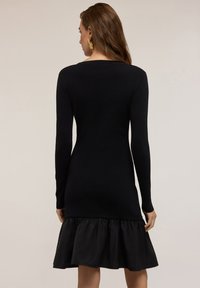 Abito nero in maglia con maniche lunghe, caratterizzato da un orlo arricciato in tessuto contrastante. Silhouette semplice e aderente con scollatura rotonda.