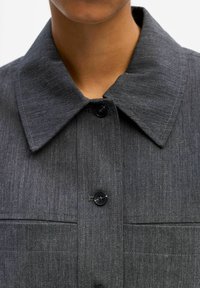 Graue Denim-Hemd mit einem großen, flachen Kragen, Fronttaschen und schwarzen Knöpfen, mit einer glatten Textur und feinem gewebtem Muster.