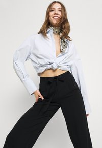 Ljusblå skjorta med knut framtill och långa ärmar, tillsammans med högmidjade svarta byxor med vida ben och en mönstrad halsduk.