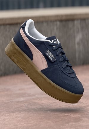 Baskets Puma Palermo bleu marine avec une bande rose, une semelle en caoutchouc et des lacets noirs, flottant au-dessus d'un sol rosé près d'escaliers en béton.