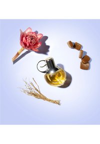 Glazen parfumfles in hartvorm met gele vloeistof, zilveren dop; roze bloem, stukjes bruine karamel en gedroogd gras ernaast op een lichtpaarse achtergrond.