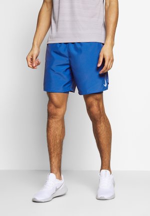 Homme portant un short de sport bleu et des baskets blanches Nike, debout sur un fond blanc uni.