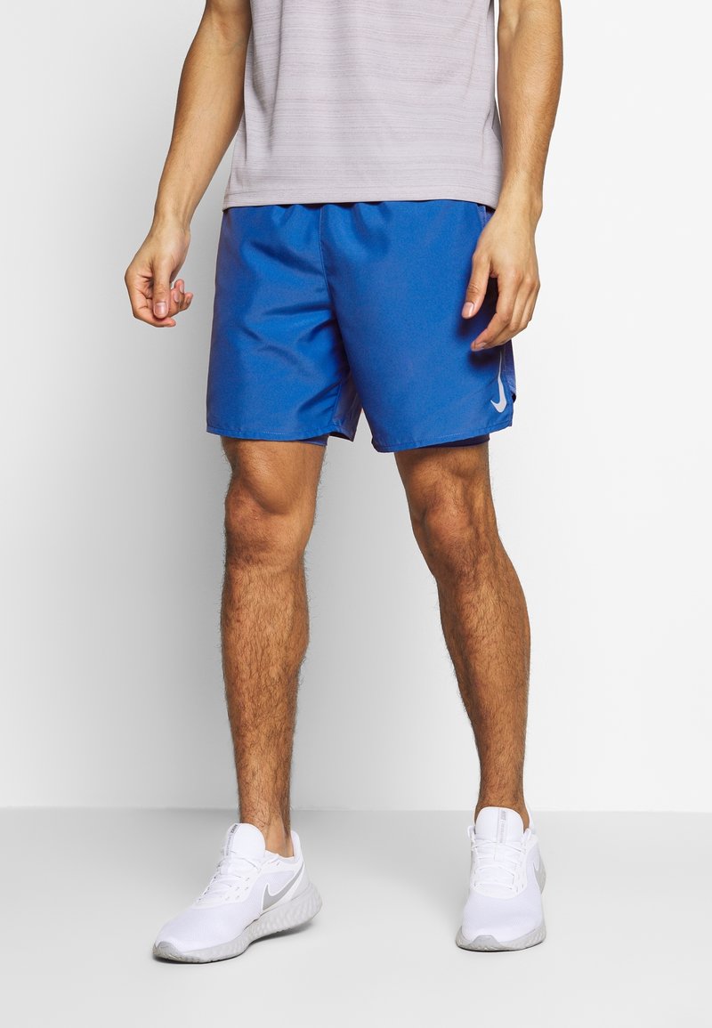 Homme portant un short de sport bleu et des baskets blanches Nike, debout sur un fond blanc uni.