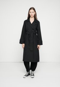 Sort trenchcoat med dobbeltradet design, brede slag og bælte med bindebånd. Kombineret med løstsiddende sorte bukser og hvide sneakers.