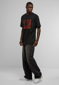 Sort bomulds T-shirt med rød grafisk tekst, "ALL DEAD," båret med mørkegrå brede jeans og hvide sneakers. Enkel, afslappet design.
