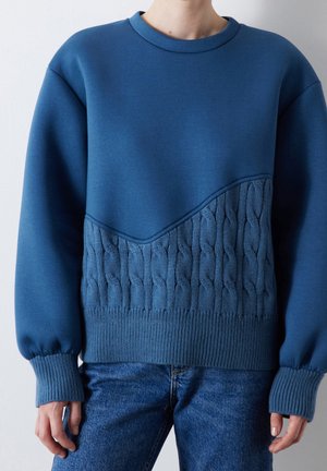 Person iført en blå sweater med en ensfarvet øverste halvdel og kabelstrik nederste halvdel, kombineret med blå denimjeans mod en hvid baggrund.