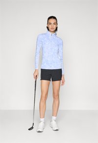 Lyseblå, langermet golfskjorte med halv-zip krage og blomstermønster, parret med svarte shorts og hvite joggesko, holder en golfklubbe.