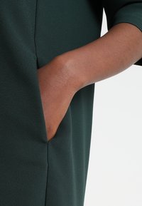 Dunkelgrüner, strukturierter Stoff mit einer Seitentasche. Nahaufnahme einer Hand, die in der Tasche ruht, mit glattem Hautton und detailreichen Merkmalen.