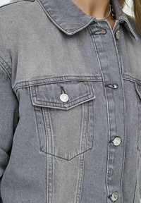 Giacca in denim grigio con chiusura a bottoni frontale, due tasche sul petto e colletto classico. Presenta dettagli in metallo argentato e una texture leggermente sbiadita.