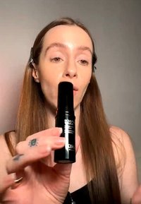 Femme aux longs cheveux bruns tient près de son visage un bâton de maquillage noir intitulé « Sculpt Touch », démontrant un produit cosmétique.