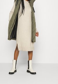 Knä-lång beige virkade klänning med sidofickor, i kombination med en olivgrön pufferväst och vita anklarstövlar med svarta sidopaneler.