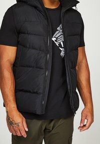 Černá péřová vesta s nastavitelnou kapucí, vybavená předním zipem a kapsami. Nositelná přes černé tričko s grafikou stříbrného tygra.