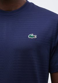 Κοντινό πλάνο σκούρου μπλε υφασμάτινου T-shirt με μικρό πράσινο λογότυπο κροκόδειλου στο αριστερό στήθος, που φοριέται από ένα άτομο.