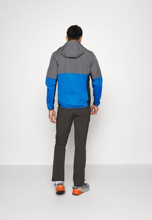 Mann mit dem Rücken zur Kamera, trägt eine grau-blaue Kapuzenjacke, schwarze Hose und graue Trailschuhe mit orangen Absätzen vor einem einfarbigen Hintergrund.