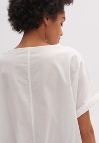 Witte linnen top met een losse pasvorm, wijde halslijn en delicate textuurafwerking aan de mouwen. Eenvoudig design met minimale details.