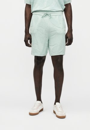 Άνδρας που φοράει ανοιχτό πράσινο σορτς από πετσέτα με σχέδιο και κορδόνι, λευκά αθλητικά παπούτσια, στέκεται σε λευκό φόντο.