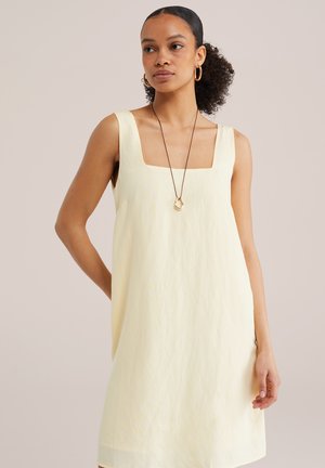 Žena s kučeravými vlasmi zviazanými dozadu nosí žlté bezrukávové šaty a zlaté kruhové náušnice s dlhým príveskom na náhrdelníku.