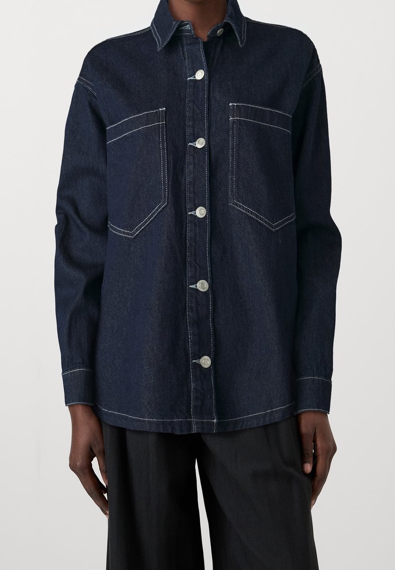 Chemise en jean bleu foncé à boutons, avec couture blanche et deux poches poitrine portée avec un pantalon noir sur une personne.