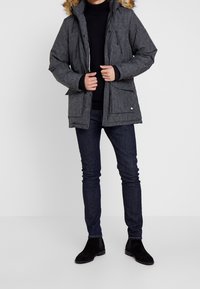 Parka acolchada gris con capucha forrada de piel, cierre delantero con cremallera y botones. Dos grandes bolsillos frontales y puños acanalados. Combinada con jeans oscuros y botas.