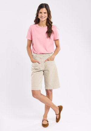Femme souriante portant un t-shirt rose, un short beige jusqu'aux genoux et des chaussures Mary Jane marron, debout les mains dans les poches et les jambes croisées.