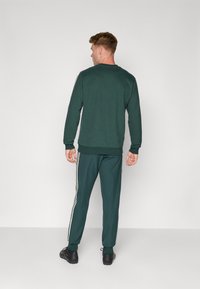 Grüner Pullover und passende Hose aus weichem Stoff. Die Hose hat weiße Seitenstreifen. Das Outfit hat eine lässige Passform mit Rippbündchen.