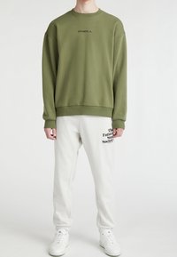 Oliwkowa bluza z okrągłym dekoltem i ściągaczami, zestawiona z białymi spodniami dresowymi z czarnym napisem "The Future Sun Society". Białe sneakersy.