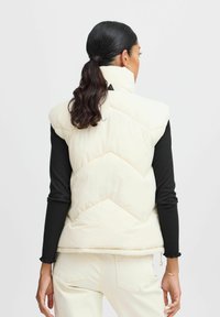 Lichtgewicht crèmekleurige puffer vest met chevronstiksels, hoge kraag, ritssluiting en zijzakken. Draagt over een zwart geribd lange mouw t-shirt.