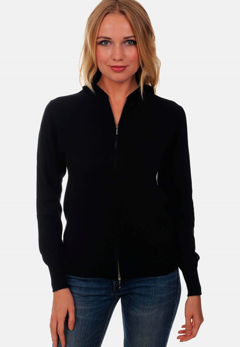 Sweatshirt noir zippé avec un col montant, fabriqué dans un matériau doux. Il présente des manches longues et une coupe ajustée, associé à un jean bleu.