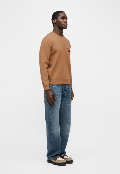 Άνδρας που φοράει καφέ πουλόβερ με σχεδιασμό κόκκινου αλεπού, φαρδιά μπλε τζιν και μπεζ-καφέ παπούτσια με κορδόνια, στέκεται μπροστά σε απλό λευκό φόντο.