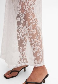 Hvid blondekjole med blomster mønstre, gennemsigtig tekstur, med en knælang skæring og sideslag. Sorte strop sandaler komplementerer looket.