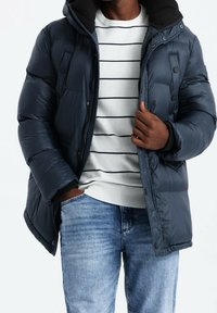 Uomo con giacca a vento blu scuro sopra un maglione bianco con strisce nere e jeans azzurri, con la mano in tasca che sistema la giacca.