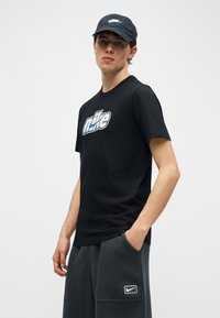 Ung mann iført svart Nike-caps, svart Nike-t-skjorte med logo og mørkegrå Nike-shorts med logo, står med en hånd i lommen.