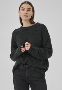 Ženska s ravnimi rjavimi lasmi, oblečena v temno sivo pleteno jopico in črne kavbojke, ki drži rokave jopice z obema rokama.