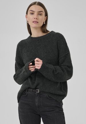 Woman with straight brown hair wearing dark gray knit sweater and black jeans, holding sweater sleeves with both hands.