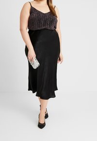 Svart, knälång kjol med en slät, satinliknande textur, tillsammans med en mörk, glittrig ärmlös topp. Håller en silverclutch och bär svarta klackar.