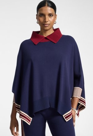 Femme aux cheveux foncés portant un poncho bleu marine avec un col rouge et des poignets rayés, assorti à un pantalon bleu marine.