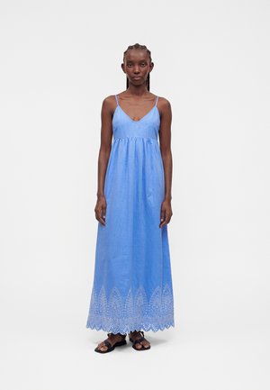 Kobieta stojąca, ubrana w niebieską, bezrękawnikową sukienkę maxi z ażurowymi koronkowymi zdobieniami na dole oraz czarne płaskie sandały.