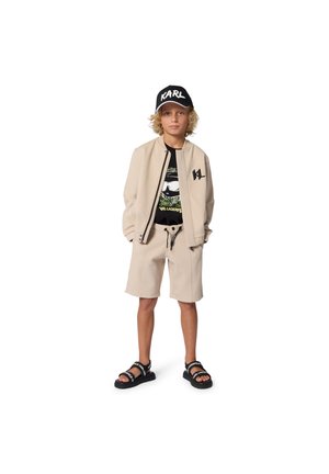 Beżowa bomberka na zamek, czarny T-shirt z nadrukiem, beżowe szorty, czarne sandały z paskami z logo oraz czapka z daszkiem w czarno-białe napisy.