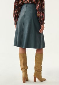 Gonna A-line in pelle verde acqua con cintura, caratterizzata da dettagli sulle cuciture. Abbinata a stivali al ginocchio in suede taupe e una blusa a fantasia.